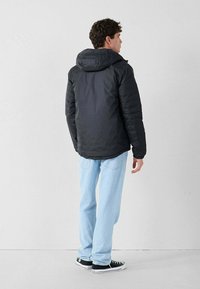 Uomo che indossa una giacca nera con cappuccio, jeans azzurri chiari e sneakers nere, in piedi di spalle contro un muro bianco semplice.