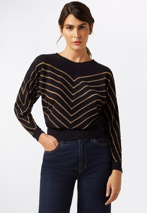 Schwarzer Pullover mit goldenen Zickzackmustern auf der Vorderseite und den Ärmeln, gerippte Bündchen und Saum, weiche Textur, lockere Passform, kombiniert mit dunklen Jeans.