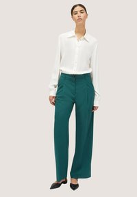 Camicia bianca con colletto alto, abbinata a pantaloni a gamba larga color teal e scarpe nere con punta; tessuto liscio e vestibilità sartoriale.
