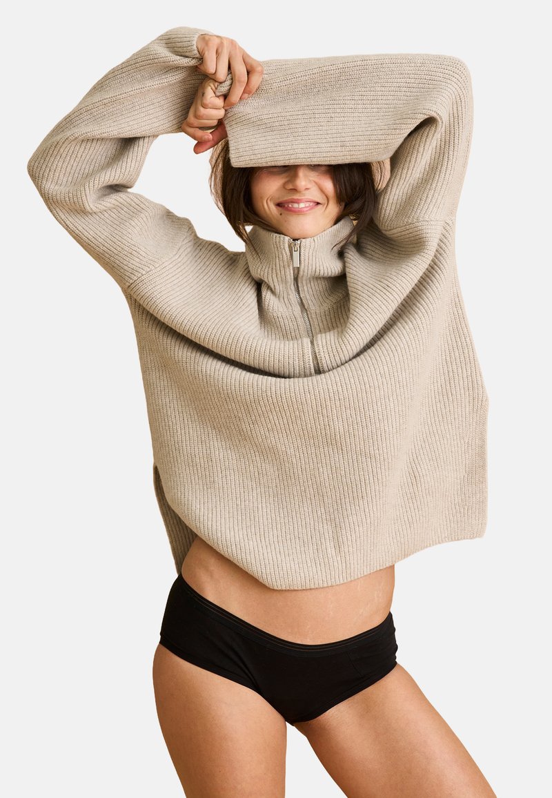 Beige ribbet sweater med høj krave og lynlåsdetalje, let oversized pasform, parret med sorte højtaljede trusser. Blød tekstur.