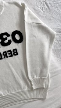 Vit stickad tröja med en iögonfallande svart grafisk tryck som säger "BERLIN" och "038." Texturerad tyg med ribbade mudd och nederkant.