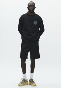 Sweat-shirt noir avec un motif graphique circulaire sur la poitrine, accompagné d'un short noir, de chaussettes blanches à la cheville et de chaussures de sport beiges.