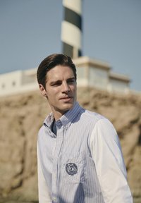 Uomo con capelli tirati indietro che indossa una camicia a righe con emblema, in piedi all'aperto vicino a un faro a strisce bianche e nere e a un terreno roccioso.