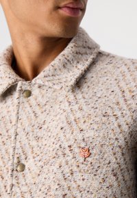 Uomo che indossa un cappotto beige testurizzato con bottoni metallici e una piccola spilla a forma di foglia d'acero rossa con l'etichetta "Scotch & Soda" sul petto.