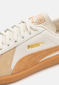 Puma sneakers with a white leather upper, beige suede accents, gold logo detail, and a textured gum rubber sole. Classic lace-up design.