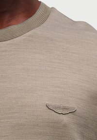 Μπλούζα με κοντά μανίκια σε μπεζ χρώμα, πλεκτή, με ριμπ γιακά και υφή. Διαθέτει γκρι κεντημένο λογότυπο Aston Martin στο μπροστινό μέρος.