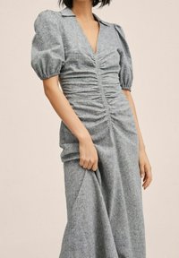 Femme portant une robe grise froncée avec des manches courtes bouffantes, un décolleté en V et un col, tenant le tissu de la jupe d'une main.