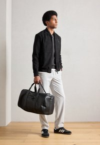Black suede bomber jacket over a grey sweater, light grey trousers, and black sneakers. Holding a large black leather duffel bag with accents.