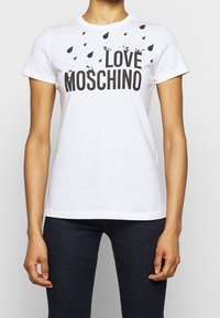 Bílé tričko s krátkým rukávem a černým nápisem "LOVE MOSCHINO" a rozptýlenou grafikou kapek deště, které má na sobě osoba v tmavých džínách.