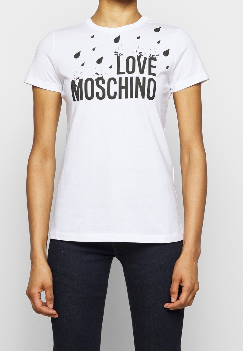 Bílé tričko s krátkým rukávem a černým nápisem "LOVE MOSCHINO" a rozptýlenou grafikou kapek deště, které má na sobě osoba v tmavých džínách.