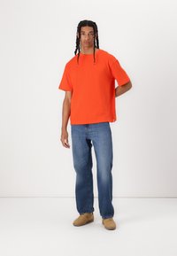 Feloranje T-shirt met korte mouwen van katoen, met een comfortabele pasvorm. Draagt het samen met blauwe jeans en tan sneakers, staand tegen een witte achtergrond.