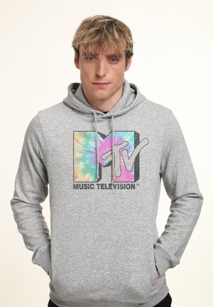 Mladý muž v šedé mikině s kapucí s barevným logem MTV Music Television s batikováním, stojící s rukama v kapsách před jednobarevným pozadím.