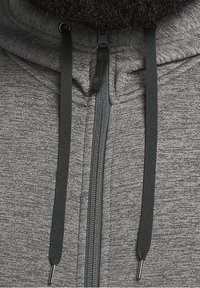 Gros plan d'un sweat à capuche gris zippé avec des cordons noirs et des embouts métalliques, montrant une partie de la barbe au-dessus du col.
