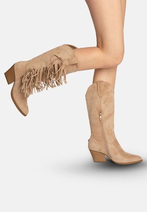 Botas beige de ante hasta la rodilla con tacones apilados, cremallera lateral y detalle de flecos en una bota, usadas con las piernas cruzadas sobre un fondo blanco.