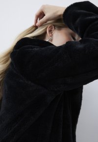 Blonde vrouw met grote kristallen oorbellen die haar haar aanpast terwijl ze een zwarte, pluizige jas draagt tegen een effen lichte achtergrond.