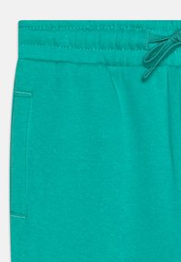 Turquoise fabric joggers with an elastic waistband and drawstring, featuring a side pocket stitched with contrasting thread.