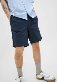 Shorts en coton bleu marine avec une coupe ample, deux poches latérales, ourlet replié, associés à des baskets blanches et des chaussettes gris clair.