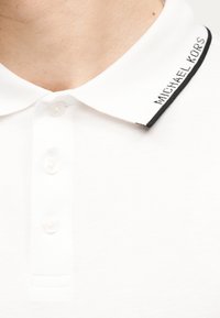 Col de polo blanc avec pointe bordée de noir et "MICHAEL KORS" brodé en noir, fermé par trois boutons blancs.
