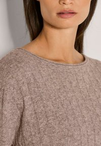 Vrouw in een beige kabelgebreide trui die de halslijn en het bovenste deel van de borst toont met steil, donker haar.