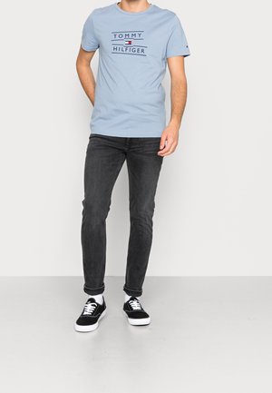 Mann trägt ein hellblaues Tommy Hilfiger T-Shirt, schwarze Slim-Fit-Jeans, weiße Socken und schwarze Sneakers und steht vor einem einfarbigen Hintergrund.