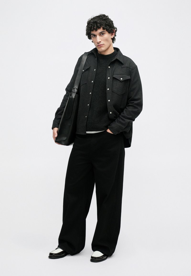 Black oversized shirt with snap buttons, layered over a dark sweater. Paired with loose black pants and two-tone shoes. Black bag slung over shoulder.