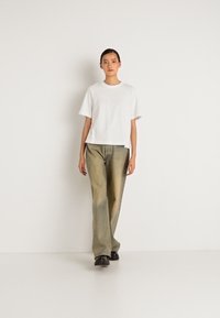 Young woman walking forward wearing a white short-sleeve shirt, olive green wide-leg pants, and black shoes against a plain white background.