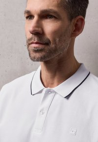 Gros plan d'un homme avec une légère barbe de quelques jours portant un polo blanc à col noir, sur un fond uni.