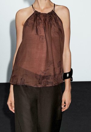 Femme portant un top dos nu marron foncé transparent avec des motifs floraux, un pantalon large sombre et un bracelet noir épais à son poignet droit.