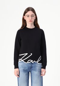 Čierna mikina s dlhými rukávmi a okr�úhlym výstrihom, s veľkým bielym kurzívnym logom "Karl" na spodnej strane. Nosená cez svetlomodré džínsy.
