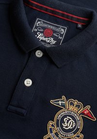 Marineblå polo skjorte med hvide knapper, rød kravekant, SuperDry-mærke og broderet University Rowing Club-logo med flag og krone.