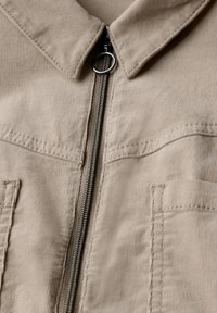 Vaaleanbeige corduroy-takki, jossa on vetoketjulla varustettu etuosa, kaulus ja kaksi etutaskua. Takissa on teksturoitu kangas ja näkyvät ompeluyksityiskohdat.