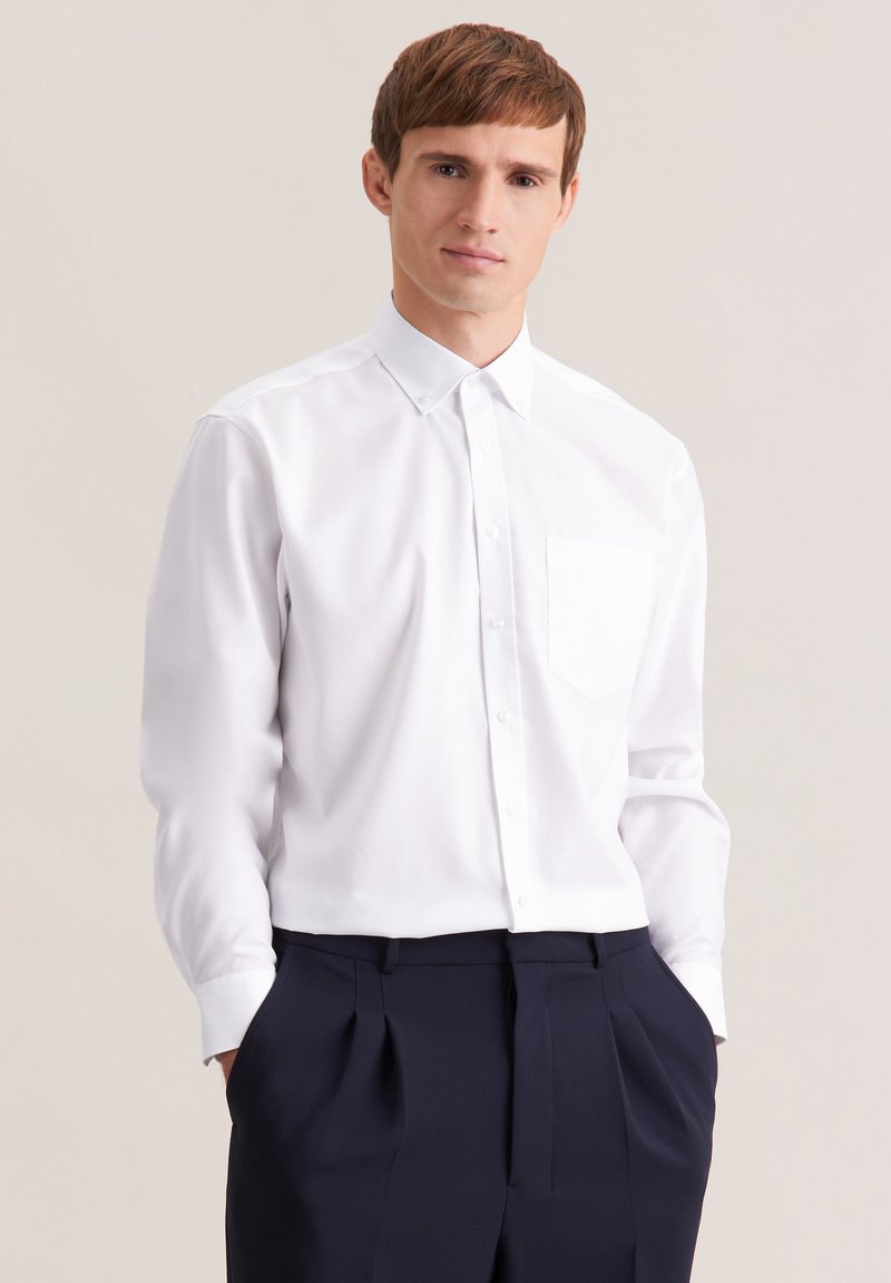 Biała koszula z długim rękawem i kołnierzykiem na guziki, z kieszonką na piersi, w zestawieniu z ciemnogranatowymi spodniami. Gładka tkanina i dopasowany krój.
