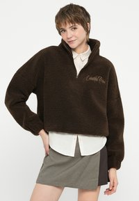 Pulover din fleece maro cu fermoar de un sfert, design scurtat și text brodat. Purtați peste o cămașă albă cu guler și o fustă cu carouri.