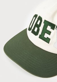 Biela a zelená čiapka s vyšitým nápisom "OBEY" v tmavozelených písmenách, má štruktúrovanú korunu a plochý zelený šilt s viditeľným šitím.