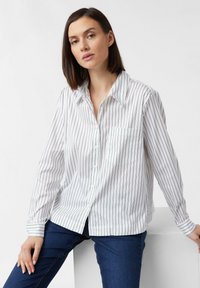 White long-sleeve shirt with black vertical stripes, classic collar, button-up front, and a chest pocket, paired with dark blue jeans.