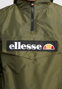 Oliven grøn jakke med halv lynlås foran, der har en fremtrædende sort rektangulær lap med "ellesse" logo i hvid og et farverigt emblem.