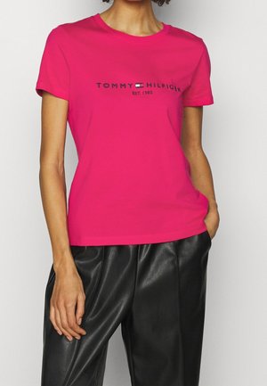 Nainen, joka pukeutunut kirkkaanpunaiseen Tommy Hilfiger -t-paitaan, jossa logo, ja mustiin nahkahousuihin, yksi käsi taskussa, valkoista taustaa vasten.