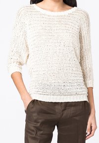 Krémový pletený svetr s volným vzorem, lodičkovým výstřihem a rukávy po tři čtvrtě. Má žebrované manžety a lem. Nošený přes hnědé kalhoty.