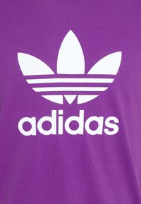 T-shirt en coton violet avec un grand logo Adidas blanc, complété par un emblème en forme de feuille à trois lobes au-dessus et trois bandes horizontales en dessous du texte.