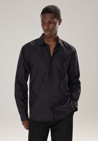 Camicia nera a maniche lunghe con tessuto floreale sottile, colletto con bottoni e bottoni frontali. Realizzata in tessuto liscio con vestibilità sartoriale.