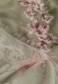 Écharpe florale en tissu léger et transparent de couleur verte, ornée de motifs de fleurs roses et marron foncé, avec des bordures à franges délicates.