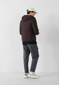 Giacca trapuntata marrone con cappuccio, caratterizzata da cuciture orizzontali. Abbinata a pantaloni cargo grigi e sneaker chiare. Cappello bianco sulla testa.