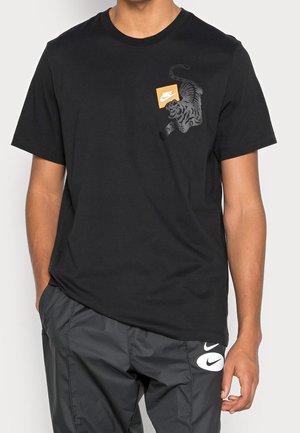Osoba nosząca czarną koszulkę Nike z grafiką tygrysa i pomarańczowym logo Nike, w połączeniu z czarnymi spodniami Nike z białymi logo Swoosh.