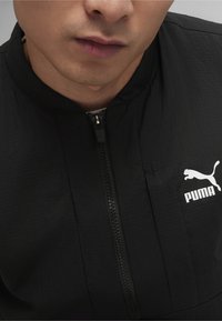 Juodas zip-up megztinis, pasižymintis tekstūruota medžiaga, stovu apykakle ir baltu Puma logotipu kairėje pusėje. Artimas apžvalga apykaklės ir užtrauktuko.
