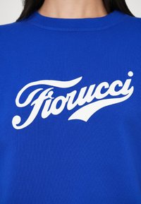 Sieviete, kas valkā zilo karaliski zilo krekliņu ar baltiem kantainiem uzrakstiem "Fiorucci" krūtīs, redzams no kakla līdz vidum.