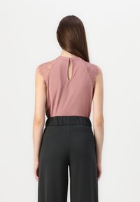 Haut sans manches en tissu doux de couleur rose, avec un empiècement en dentelle, présentant un dos en forme de clé. Assorti à un pantalon noir taille haute.