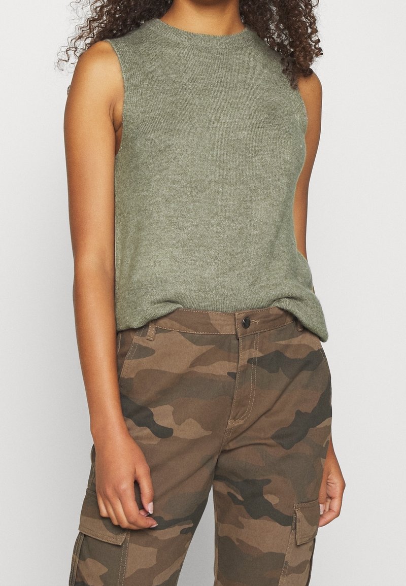 Haut tricoté sans manches vert olive associé à un pantalon cargo camouflage marron et vert porté par une femme aux cheveux bouclés.