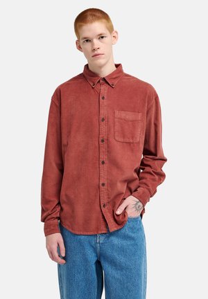Giovane uomo con capelli corti e rossi che indossa una camicia di velluto a coste color ruggine abbottonata e jeans blu, con un tatuaggio del sole sulla mano sinistra.