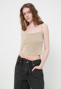 Jeune femme aux cheveux ondulés portant un crop top beige sans manches et un jean noir ample avec une taille à cordon, debout devant un fond blanc uni.