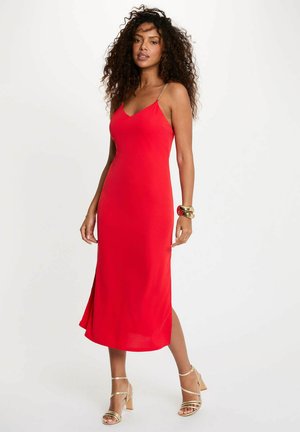 Donna con capelli ricci che indossa un vestito midi rosso senza maniche con spalline a catena dorate e sandali dorati con tacco alto su uno sfondo bianco.